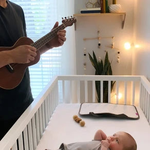 A person strumming a ukulele while a baby looks at them peacefully in a nearby cradle.