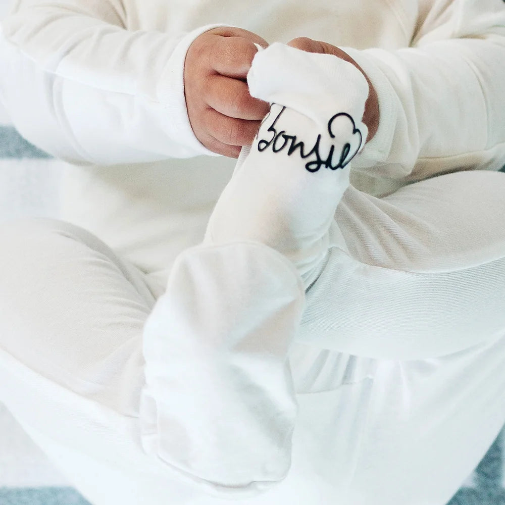 An infant wearing a white onesie by Bonsie is seated with its legs crossed. 
