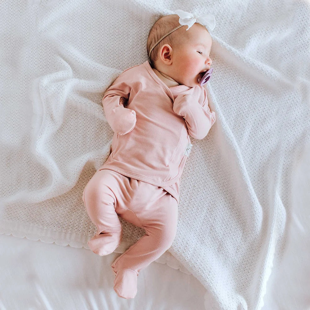 A baby wearing a light peony Bonsie footie lying down on a comfortable mattress.