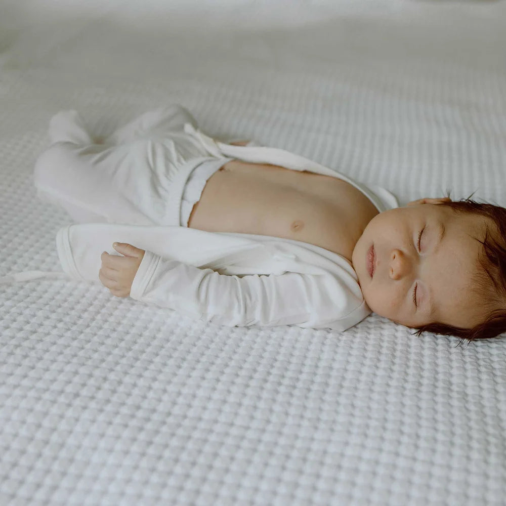 An infant sleeping peacefully on a textured white mattress wearing a white long-sleeved footie with the belly exposed.
